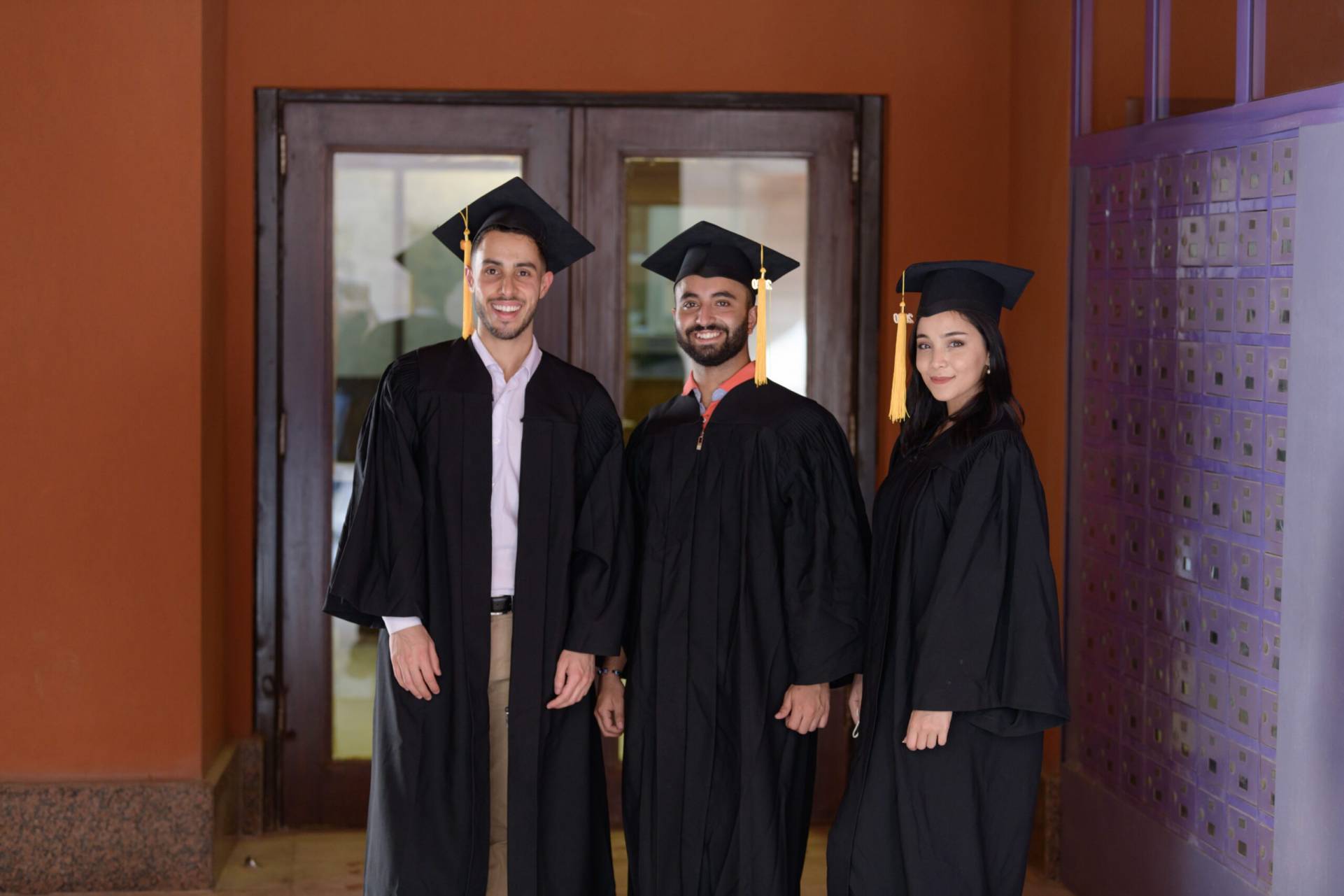 Two male students and one female student dressed in graduation attire smiling.